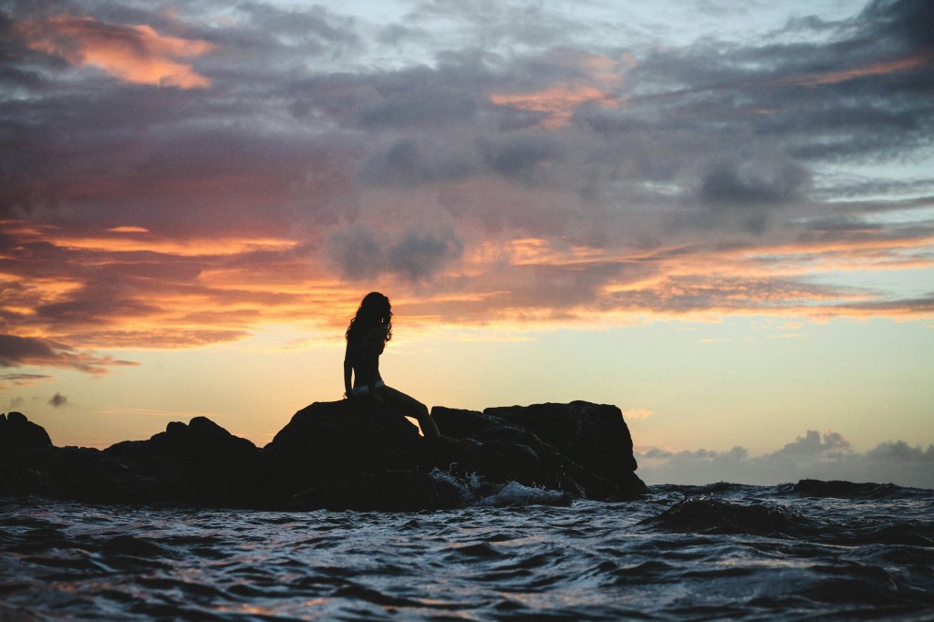 Silhouette of person sitting on rocks against a vibrant sunset sky over the ocean.
