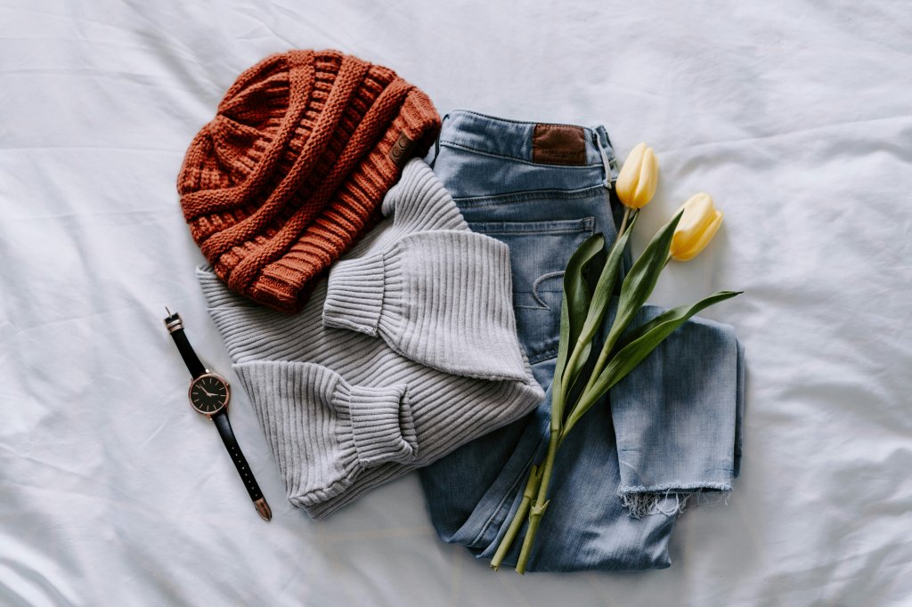 Sweater, hat, jeans, watch, and yellow tulips on a white bed.