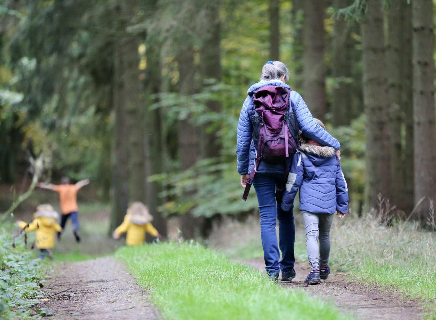 Woman and child walking on a forest path, children playing ahead.