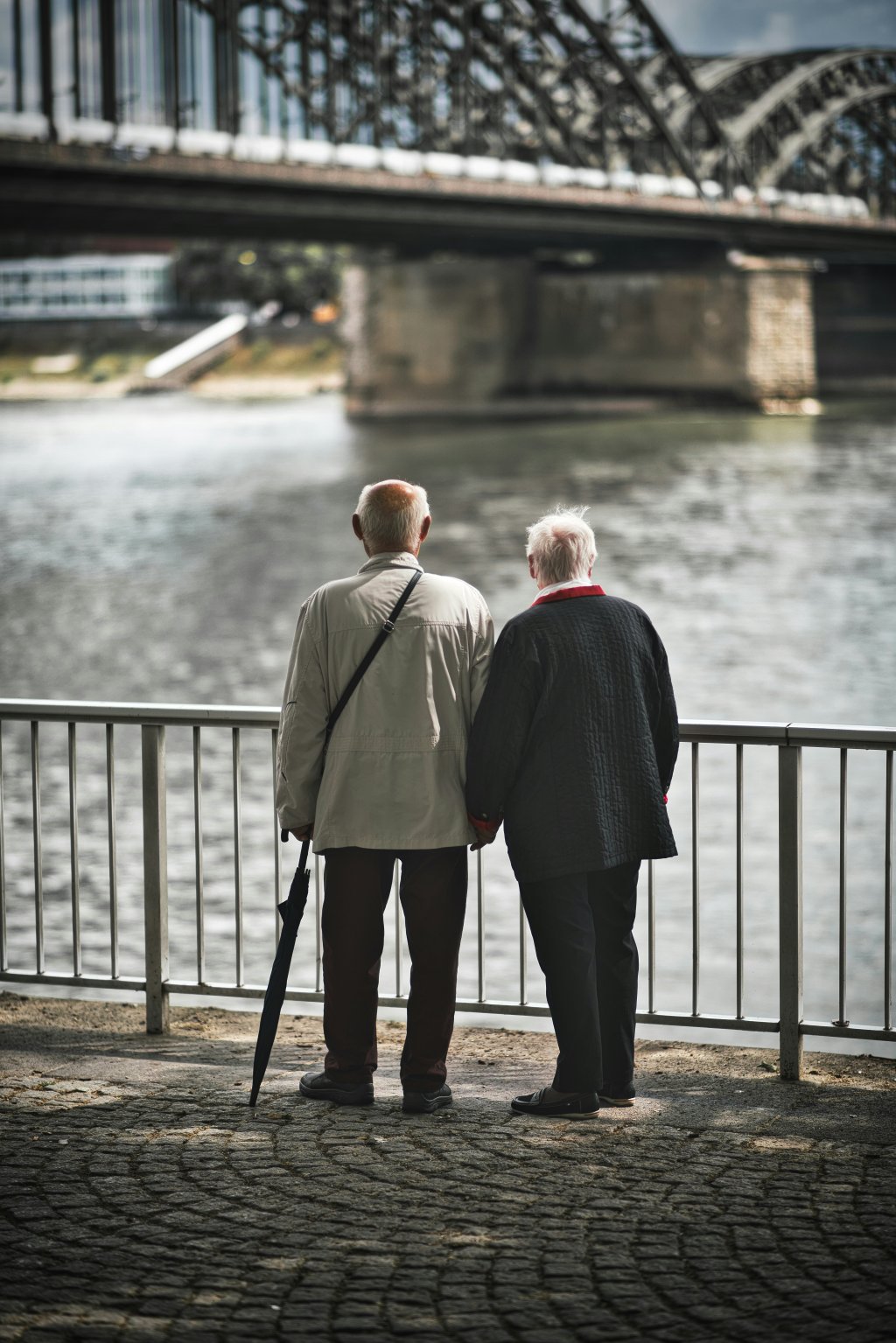 Elderly couple stands by river, holding hands, near a large bridge.