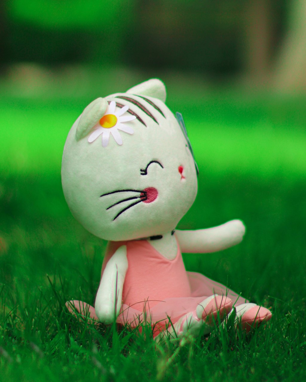 Stuffed cat toy with daisy on its head sits in grass, wearing a pink dress, smiling.