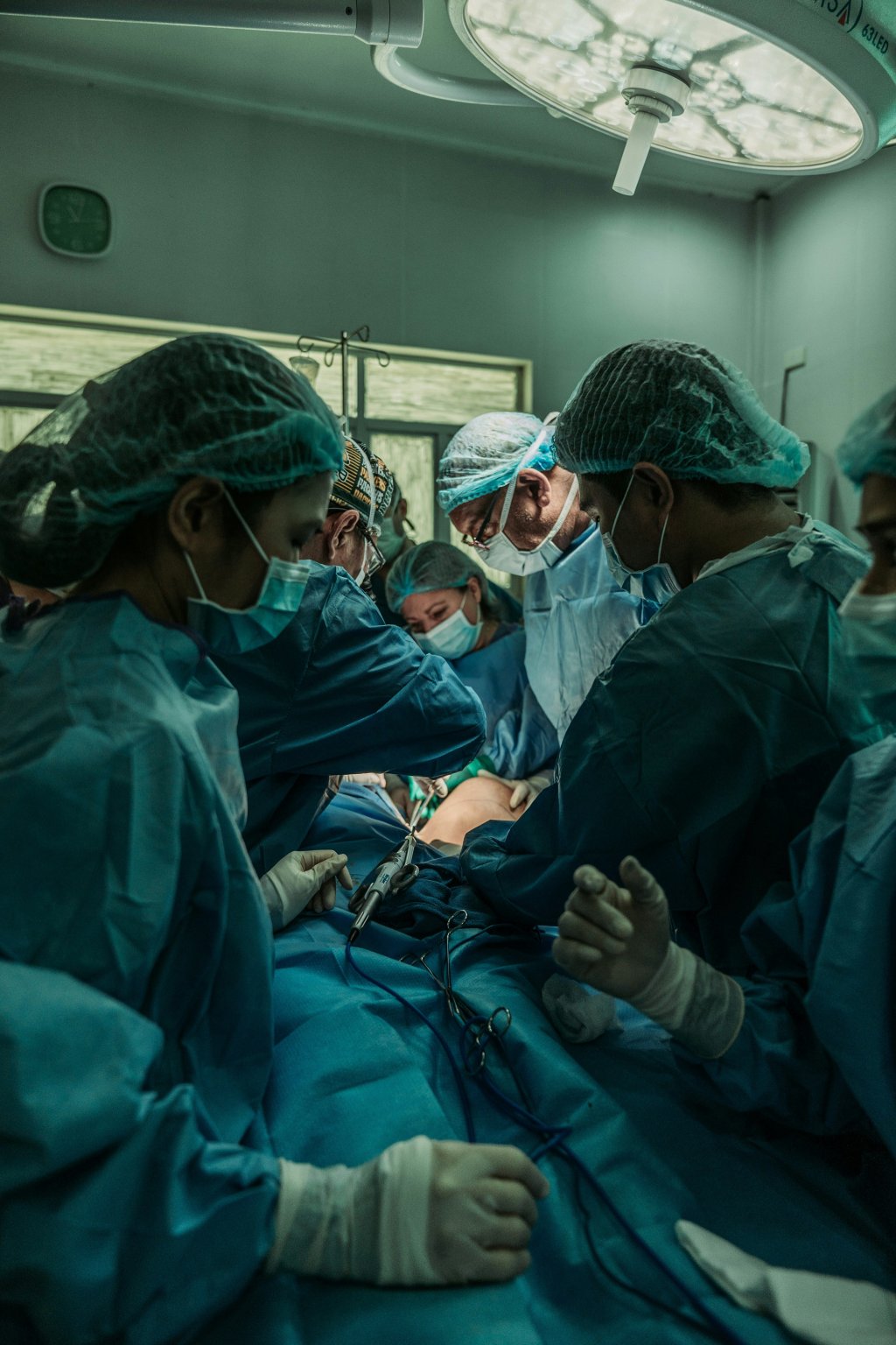 Surgical team operating in a hospital under bright light, wearing green scrubs and masks.