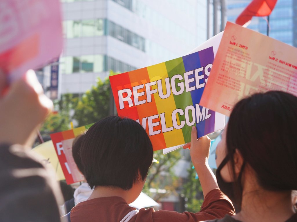 People holding a sign reading "Refugees Welcome" with rainbow colors at a gathering.