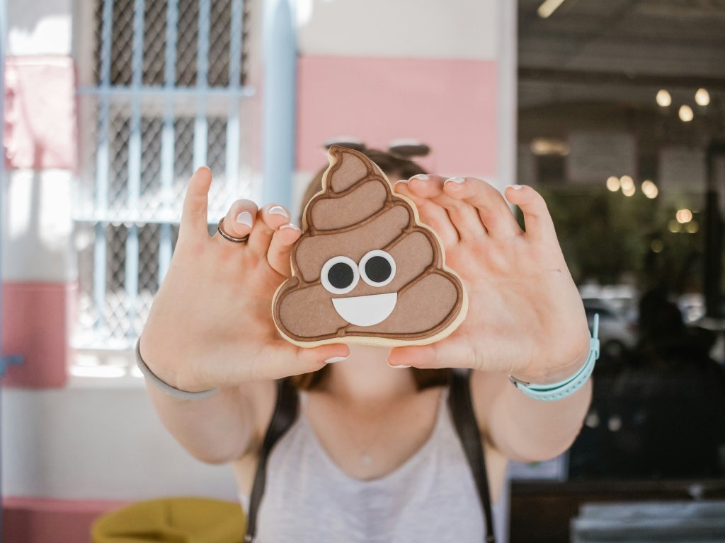 Person holding a poop emoji cookie in front of their face, outdoor setting.