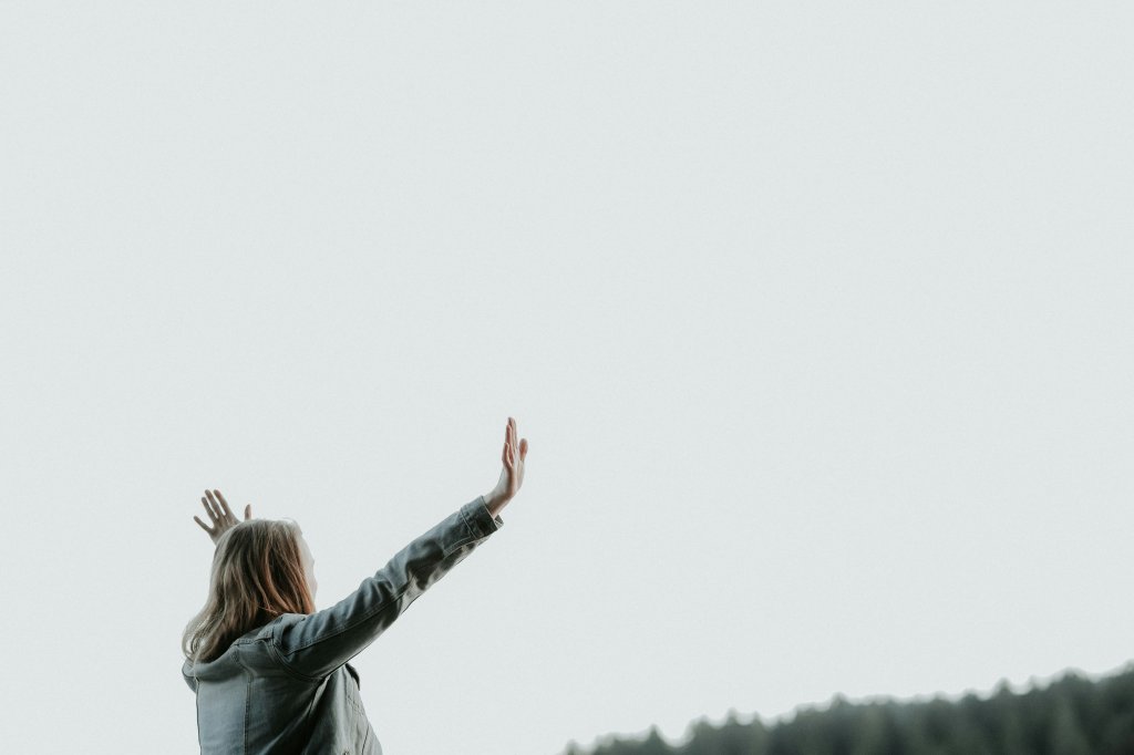 Person in jacket raises hands to the sky, with trees and a pale sky in the background.