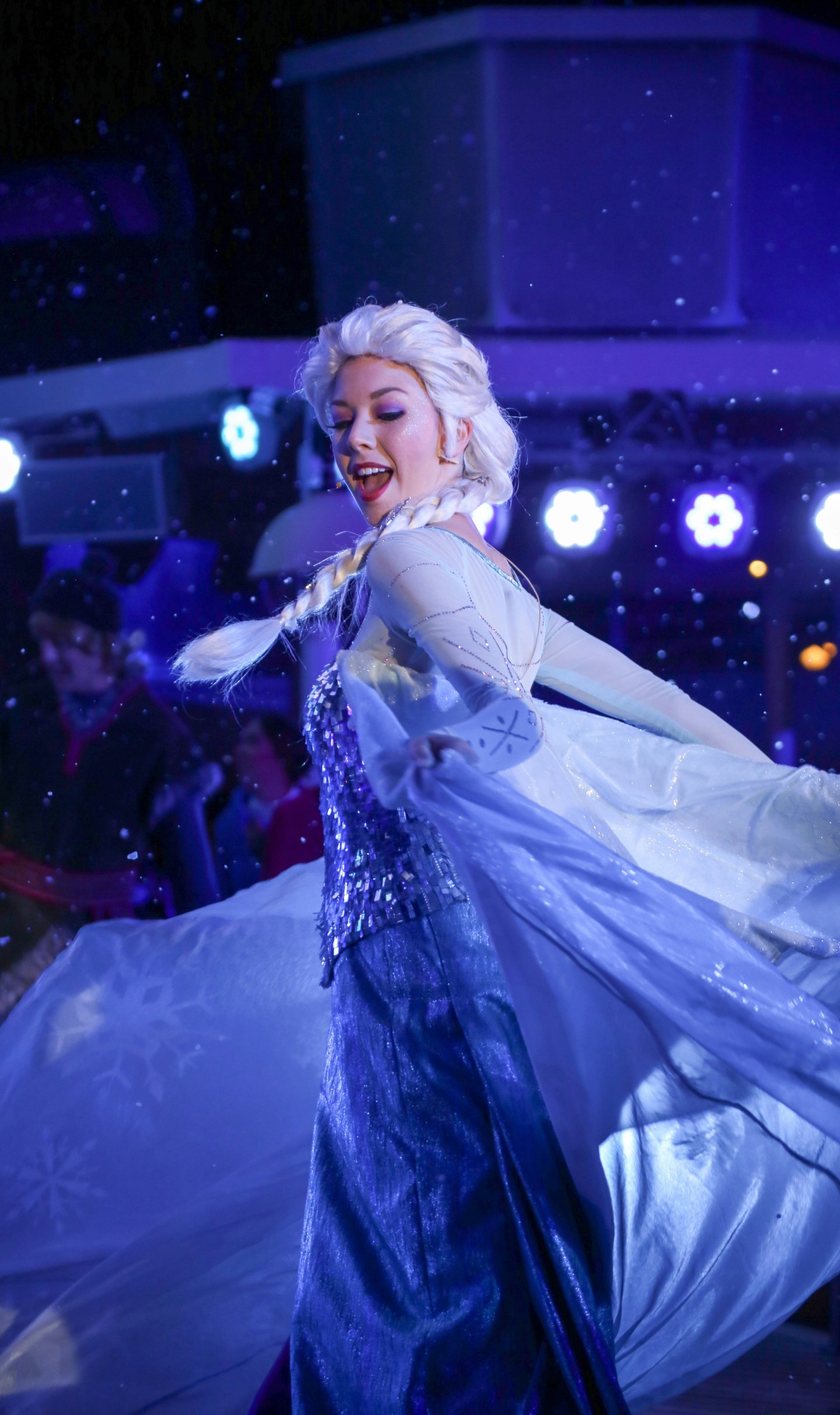 Elsa in a sparkly blue costume and white wig, dancing in falling snow.