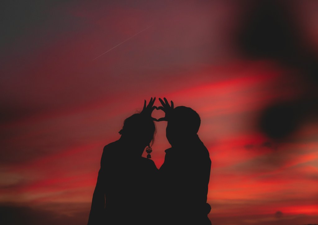Silhouetted couple forms heart with sunset sky.