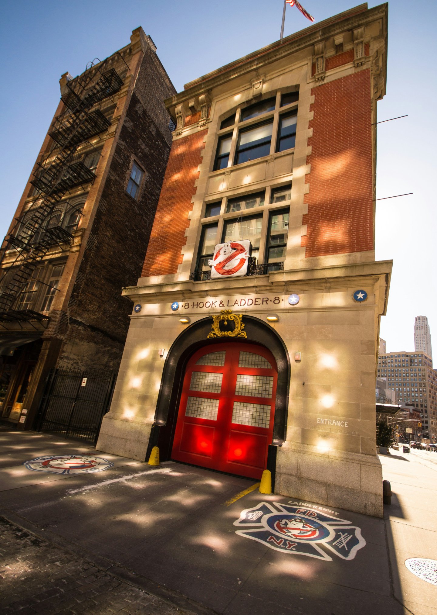 Firehouse with red doors and Ghostbusters logo, sunlight on brick facade.