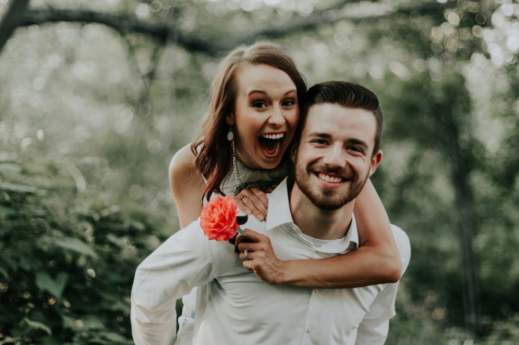 Smiling woman on man's back in forest, holding a red flower.