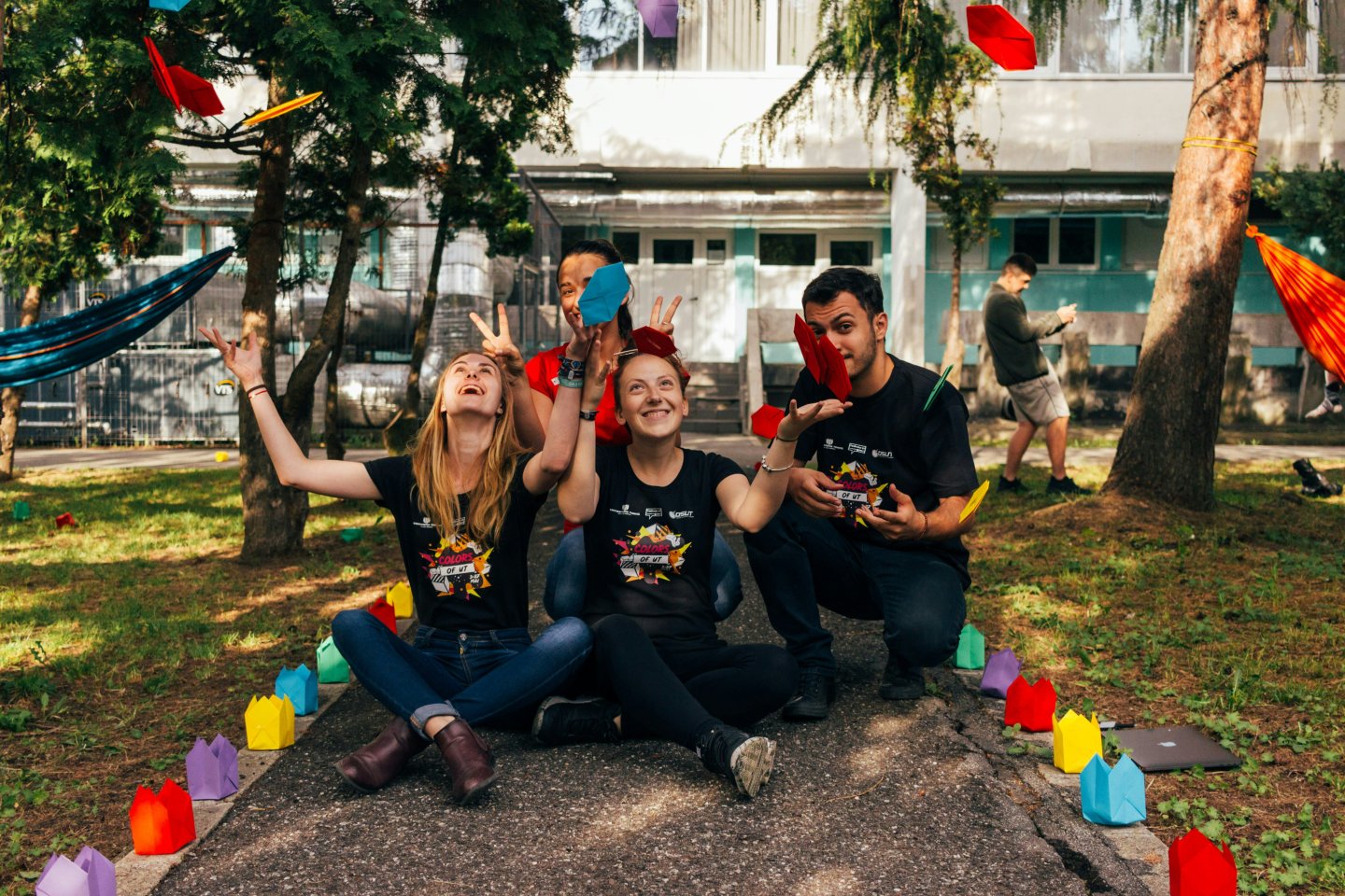 people joyfully throw paper planes outdoors, surrounded by colorful decorations.