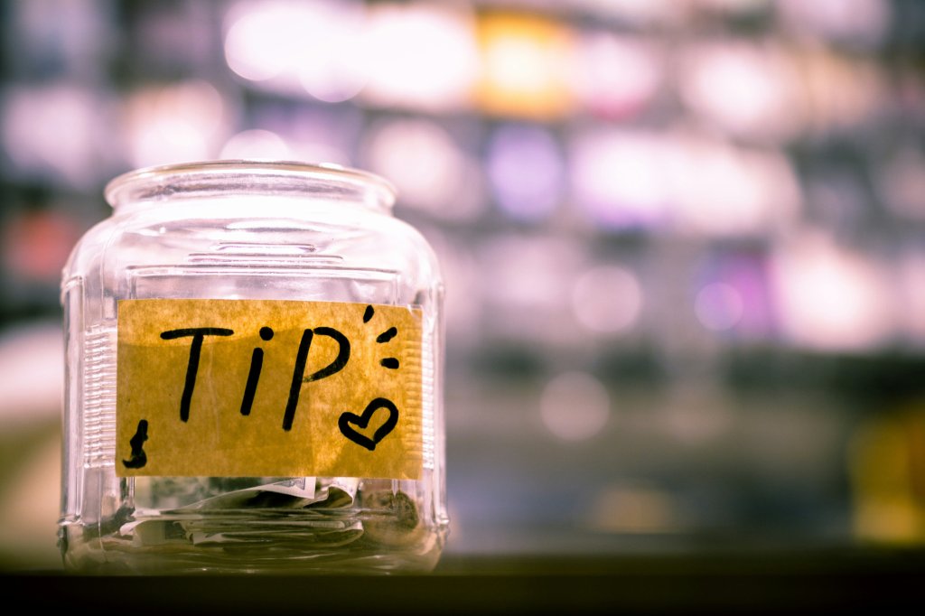 Glass tip jar with a handwritten label, blurred colorful background.
