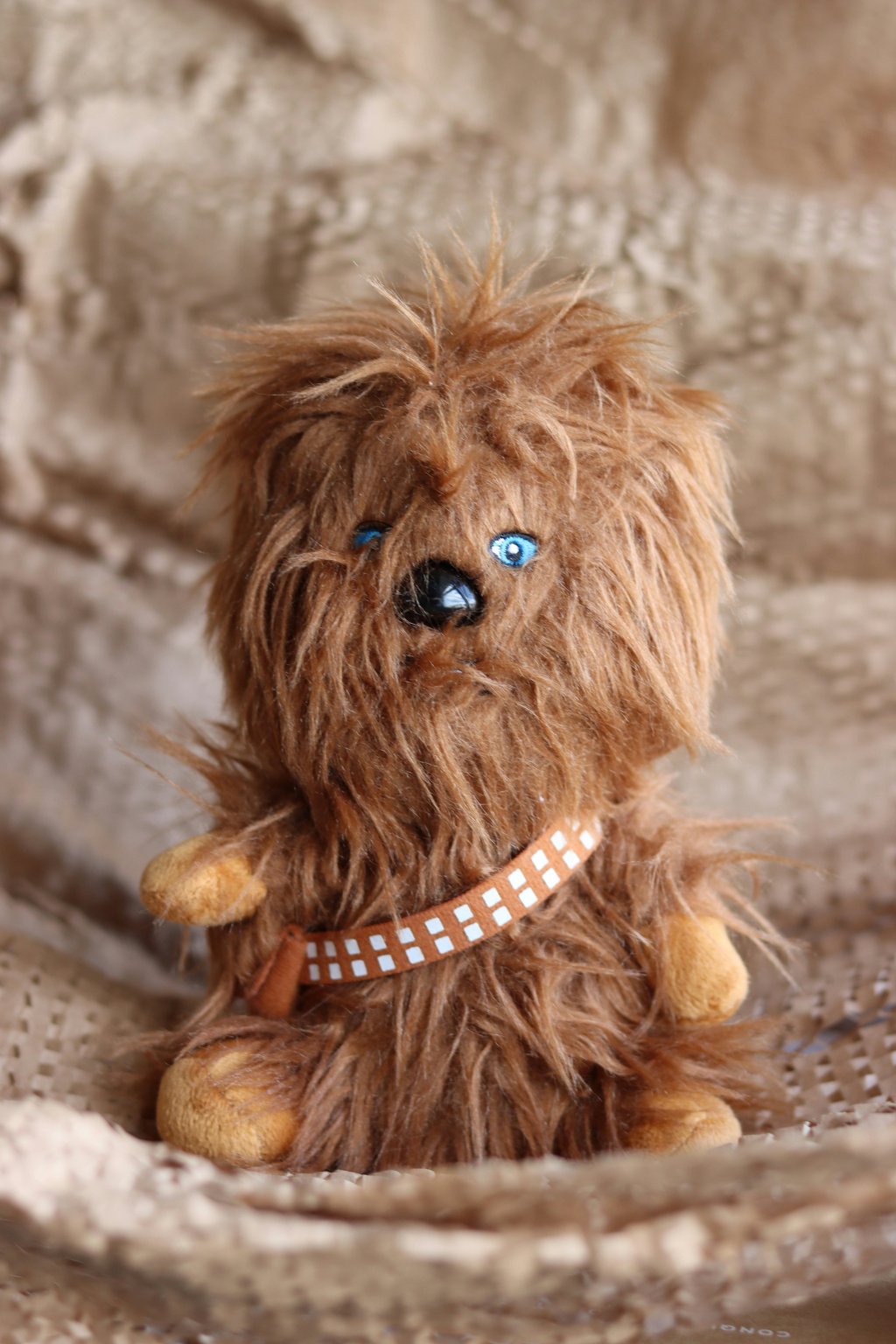 Fluffy brown stuffed toy with blue eyes, sitting on a textured beige surface.
