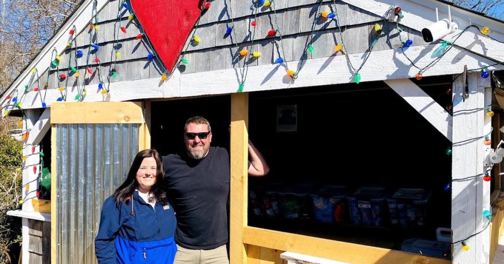 A man and woman, smiling, stand in front of a farm stand