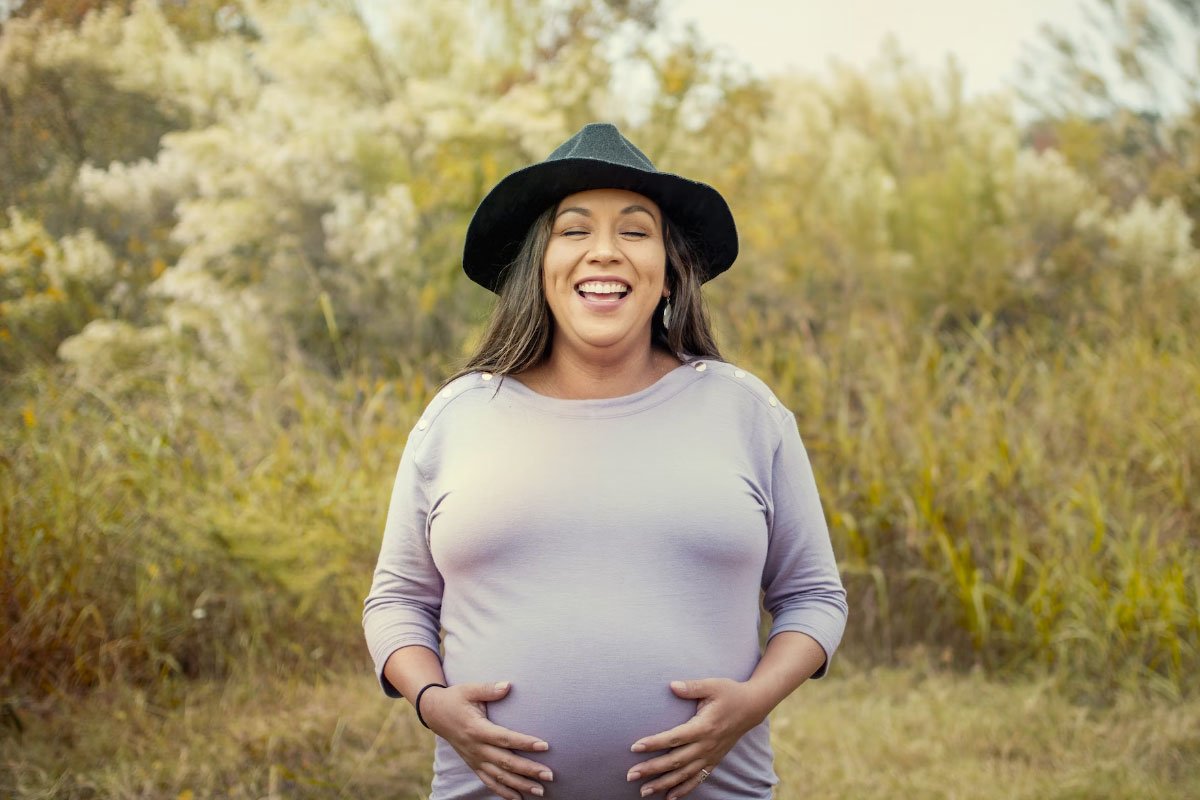 Smiling pregnant woman in a hat outdoors, touching her belly.