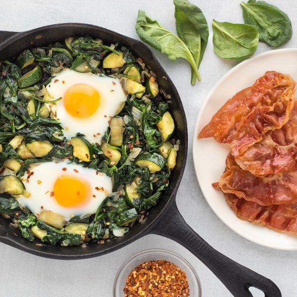 eggs and green veggies in a skillet, plate of bacon