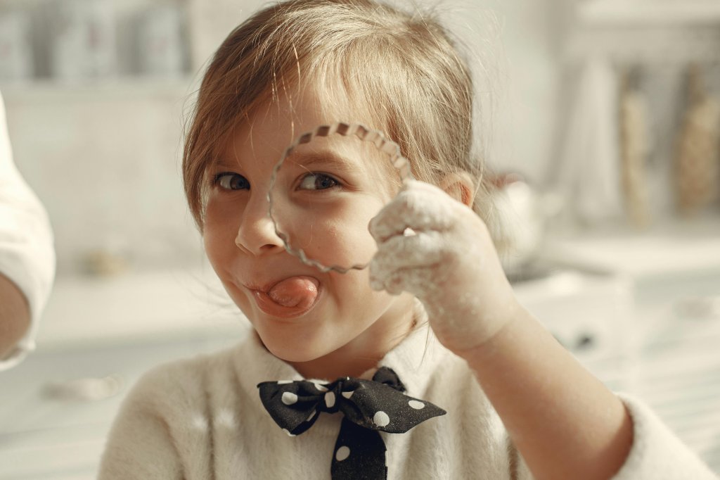 little girl with flour on her hand holding up a cookie cutter with her tongue sticking out