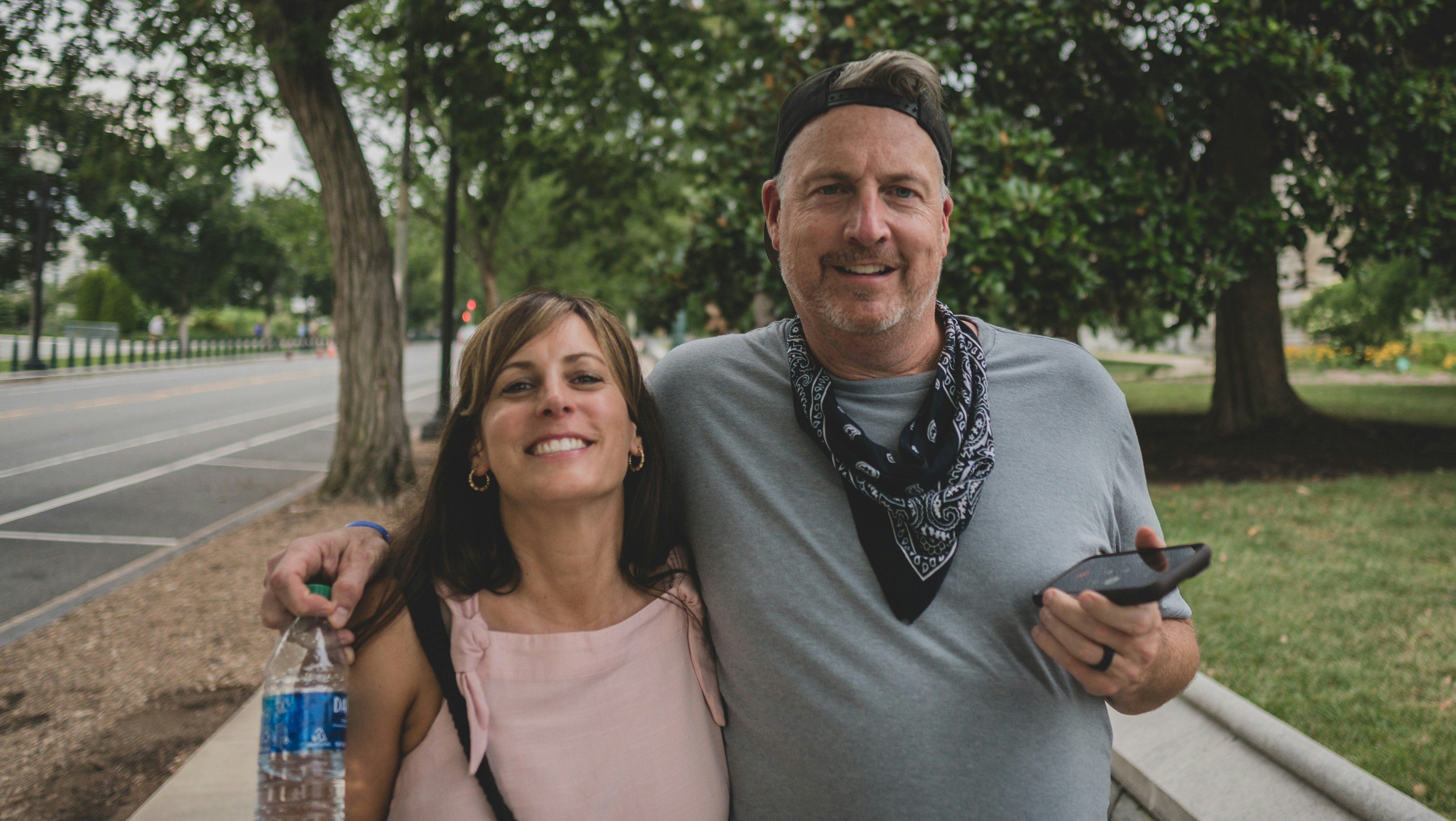 Smiling couple outdoors, woman holding water bottle, man with phone, trees in background.