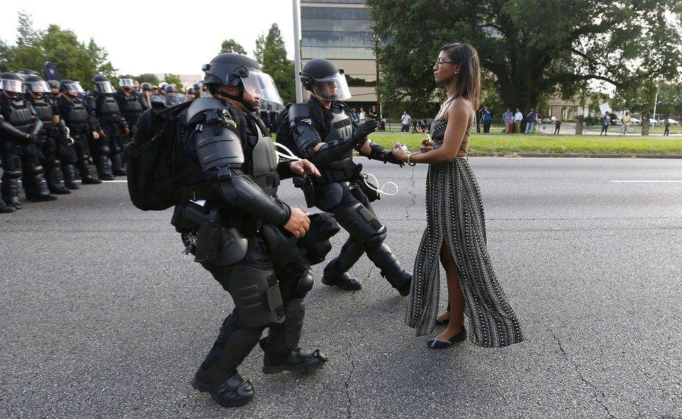 a woman stands calmly before two police officers in riot gear.