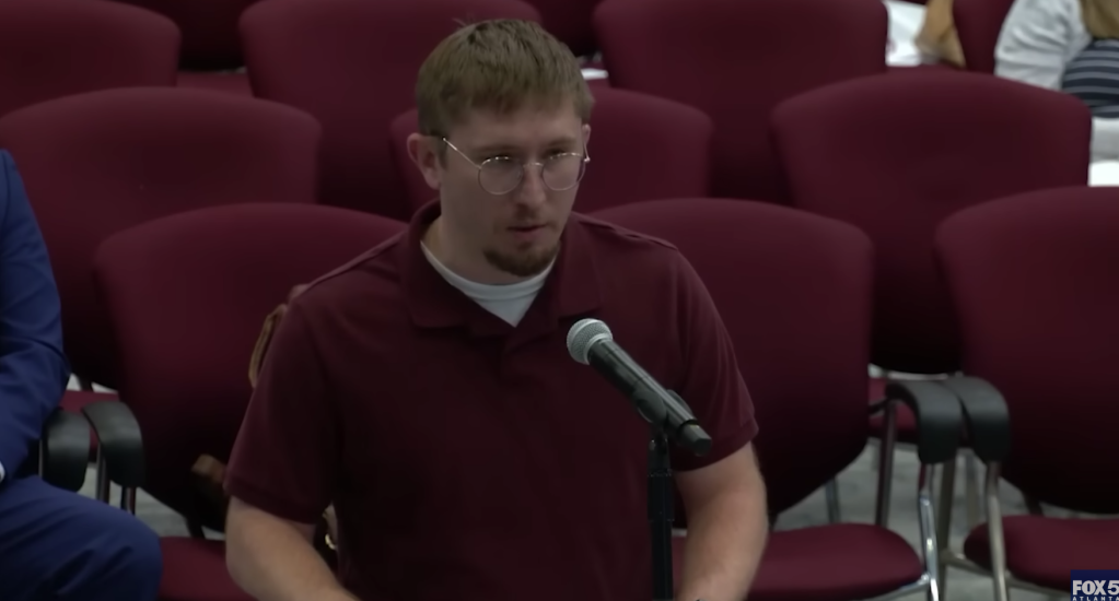 Man in maroon shirt speaking into a microphone at a public meeting.