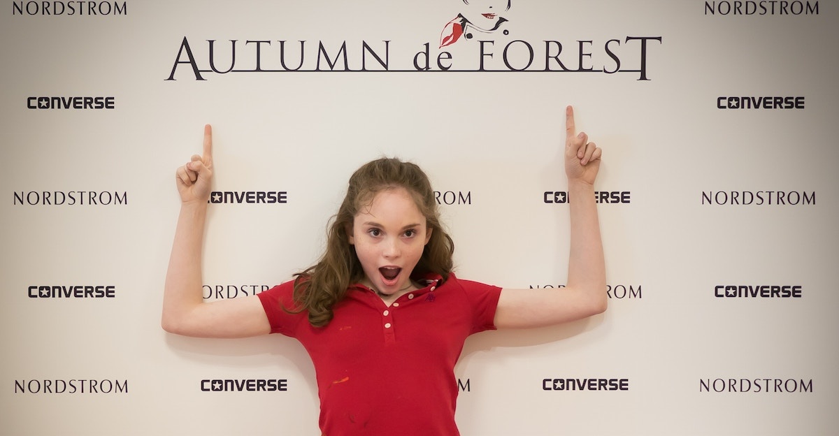 Girl in red shirt pointing up at "Autumn de Forest" sign with surprise.