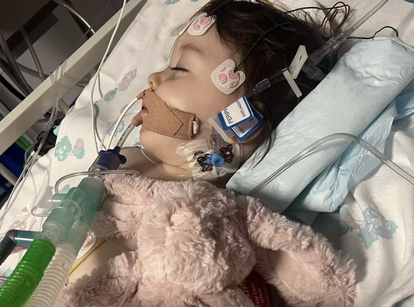 Baby girl in hospital bed with a teddy bear and oxygen mask.