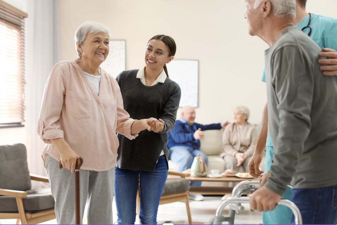 woman helps an elderly woman walk with a cane in a room with other elderly people socializing 