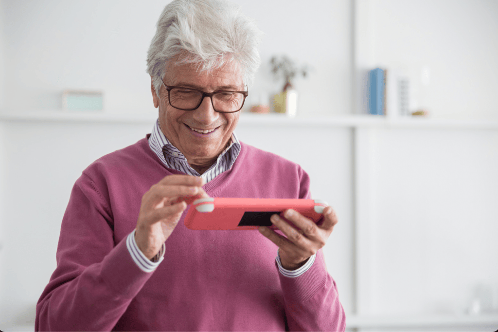 Older man in glasses and a pink sweater smiling while typing a text message on a pink smartphone held sideways.