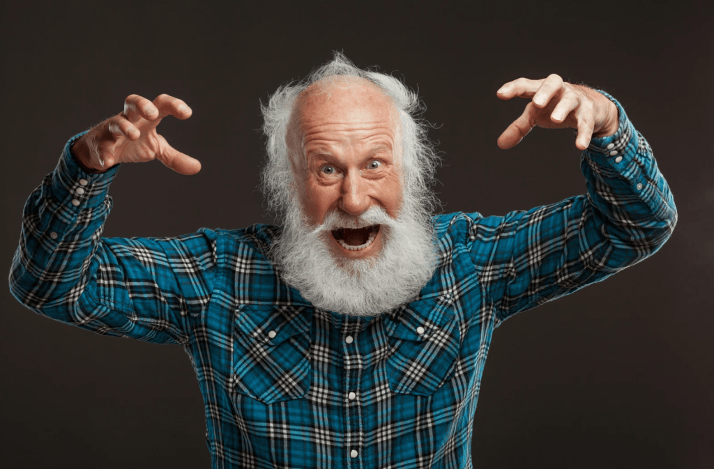 Older bearded man in a plaid shirt playfully making a spooky "boo" gesture with raised, clawed hands and a wide-eyed grin.