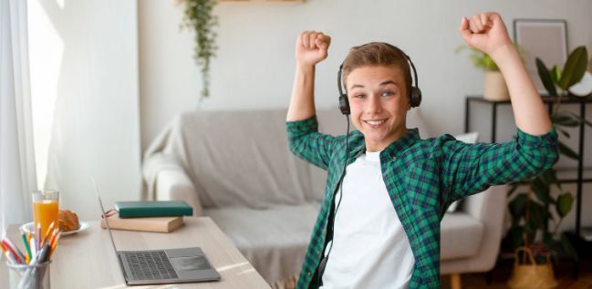 happy tween boy, happy kid, kid with arms raised, headphones, laptop, green shirt