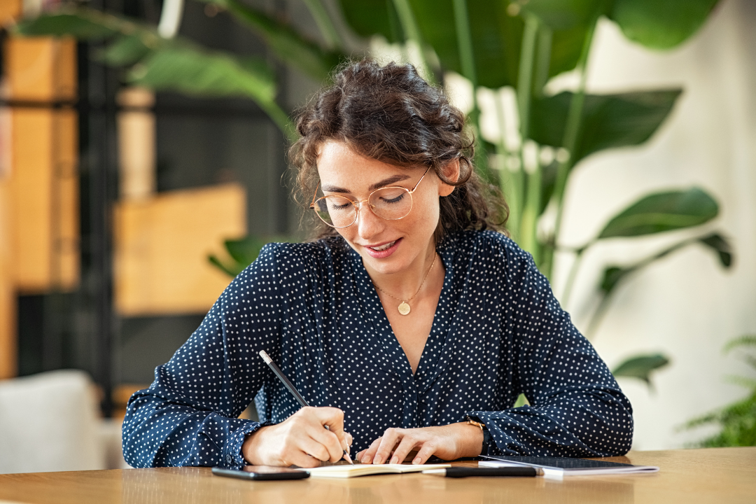 Woman in glasses writing at a desk, surrounded by plants.