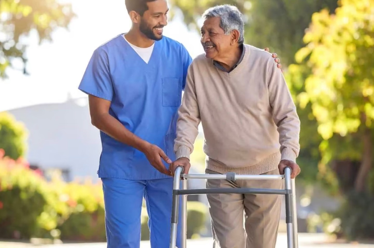 A caregiver in medical scrubs helps an older man with a walker.