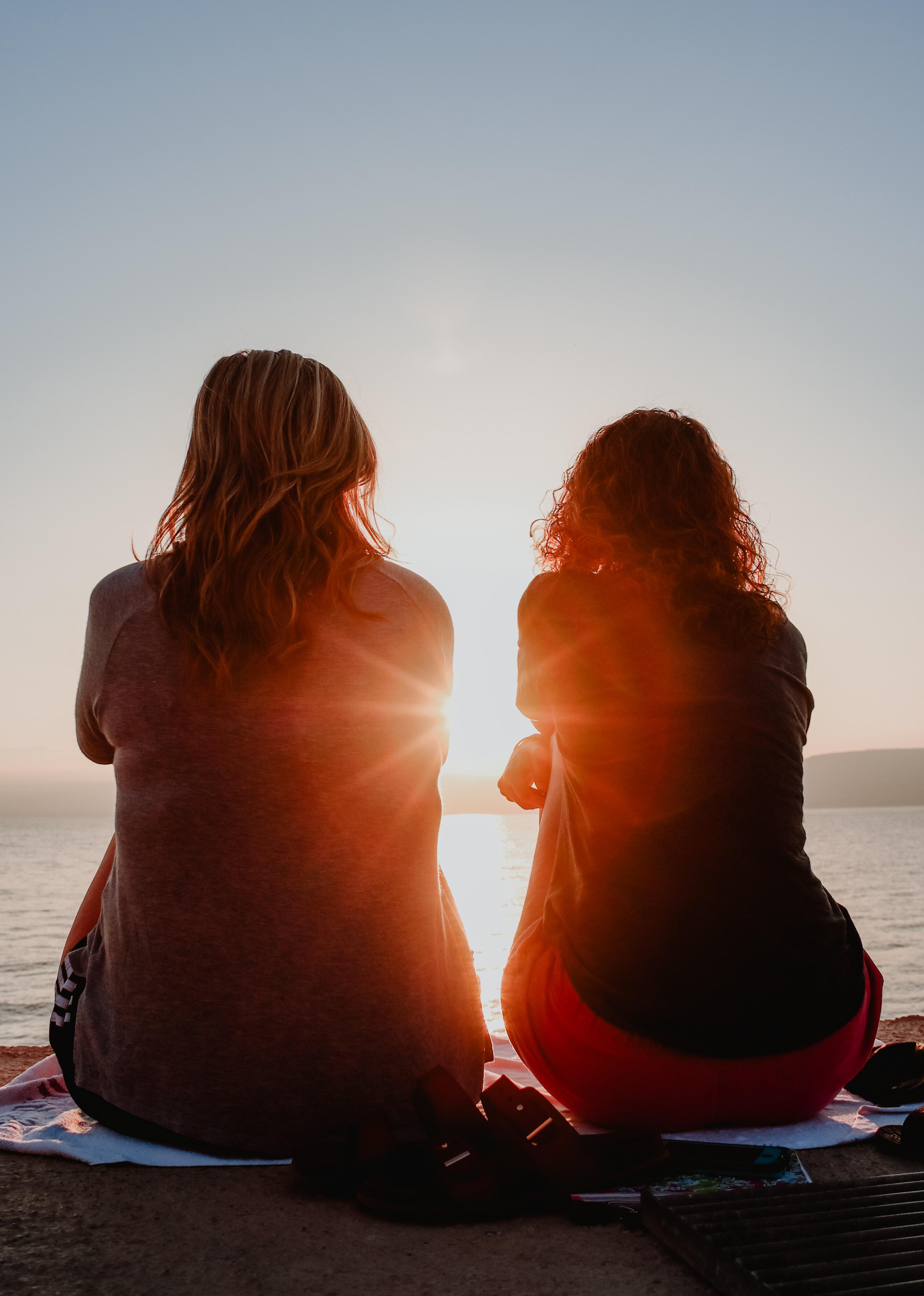 two women sitting on beach facing ocean sunset