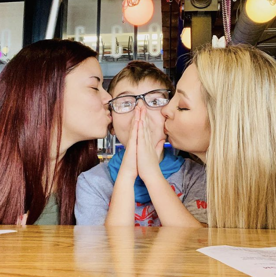 two women kissing a little boy wearing glasses on his cheeks