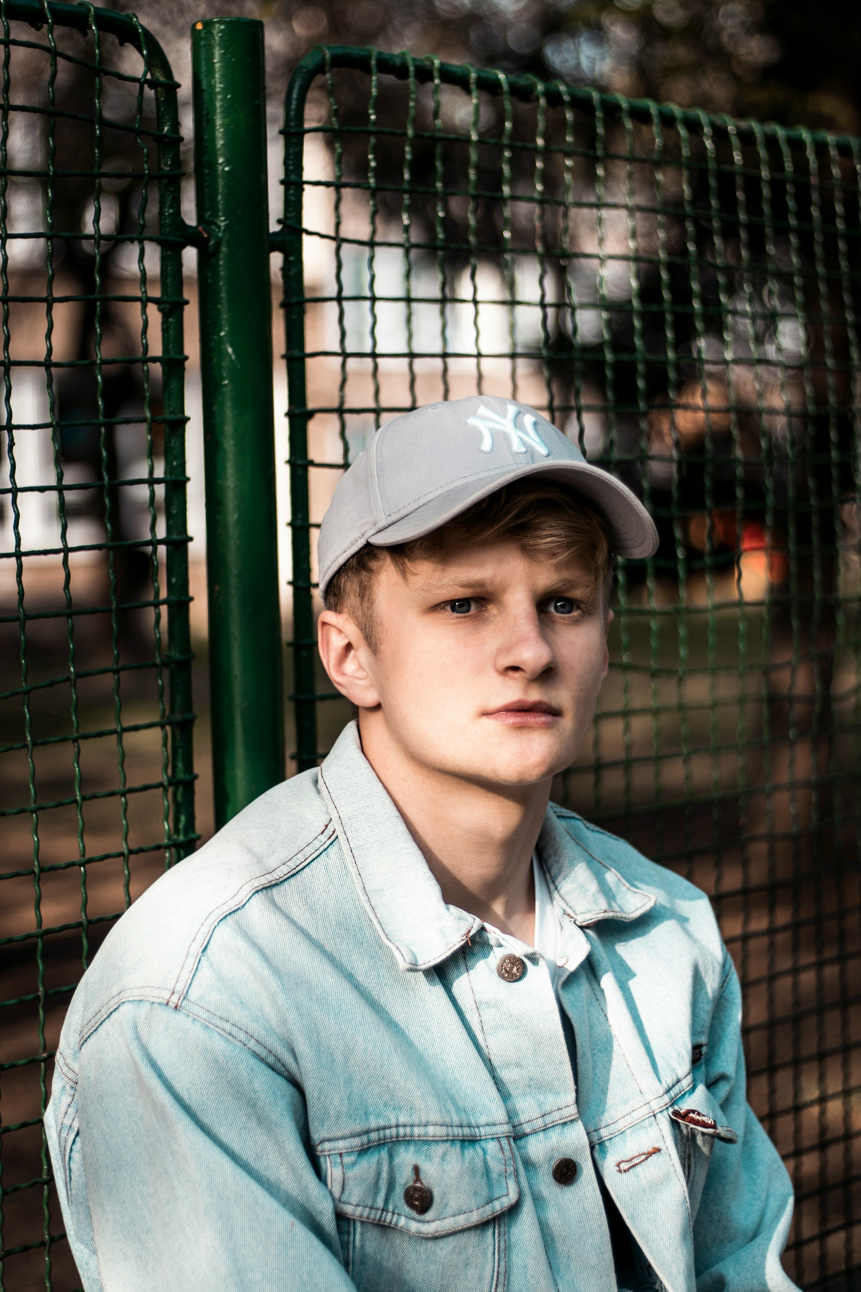 boy wearing blue button-up denim jacket sitting beside green fence