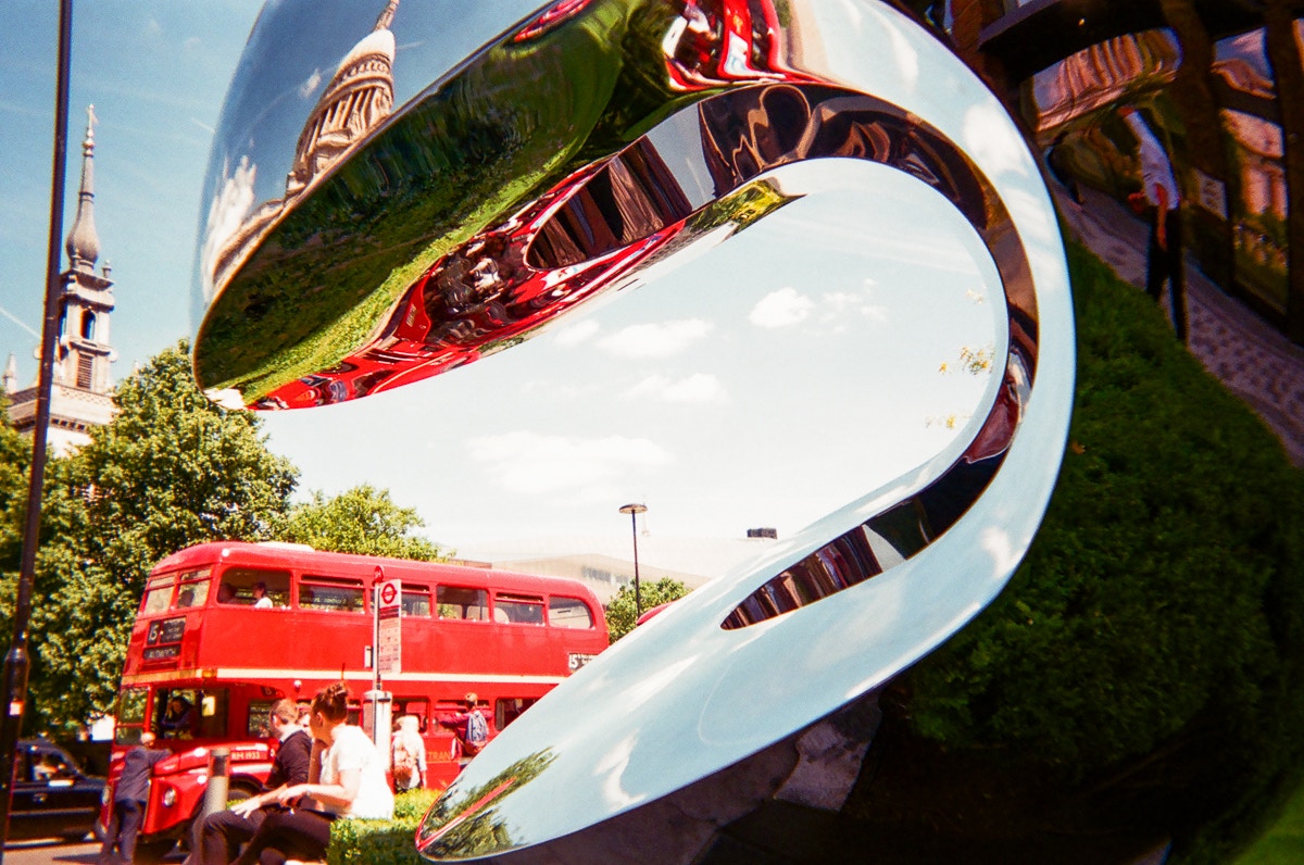 A red-double-decker-bus behind a mirrored sculpture.