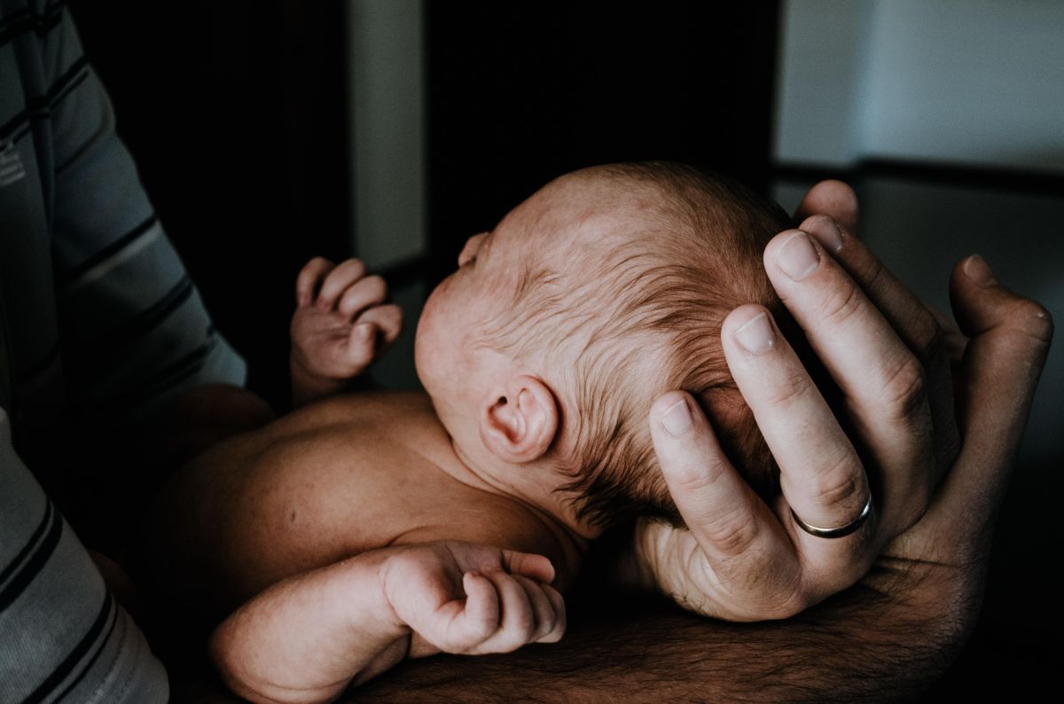 Man's hand with gold wedding band holding a newborn baby's head.