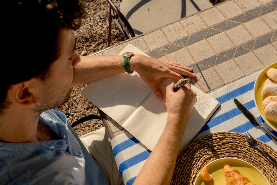 A man sits at an outdoor table writing in a journal.