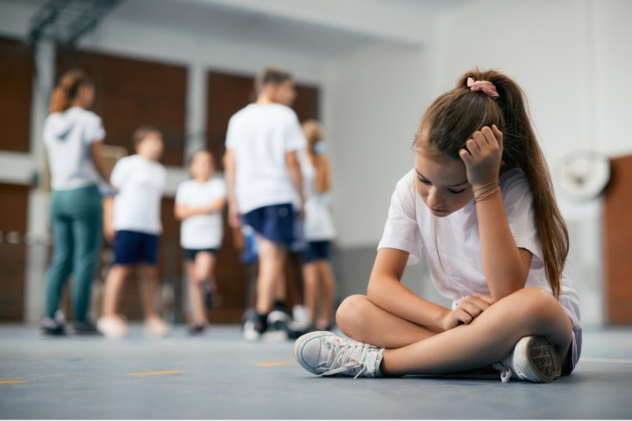 A girl sits away from the group, appearing to be left out in gym glass