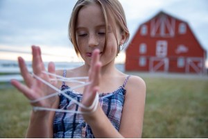 A girl stands outside near a barn doing cat's cradle with a string