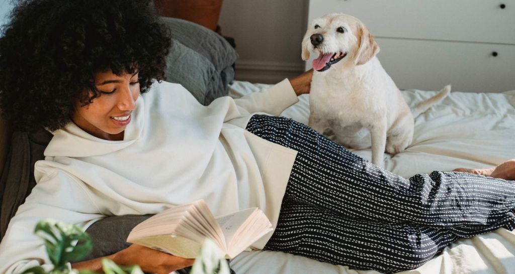 woman laying in bed reading a book with her dog, woman enjoying reading, Black woman enjoying reading while laying on her bed with her dog