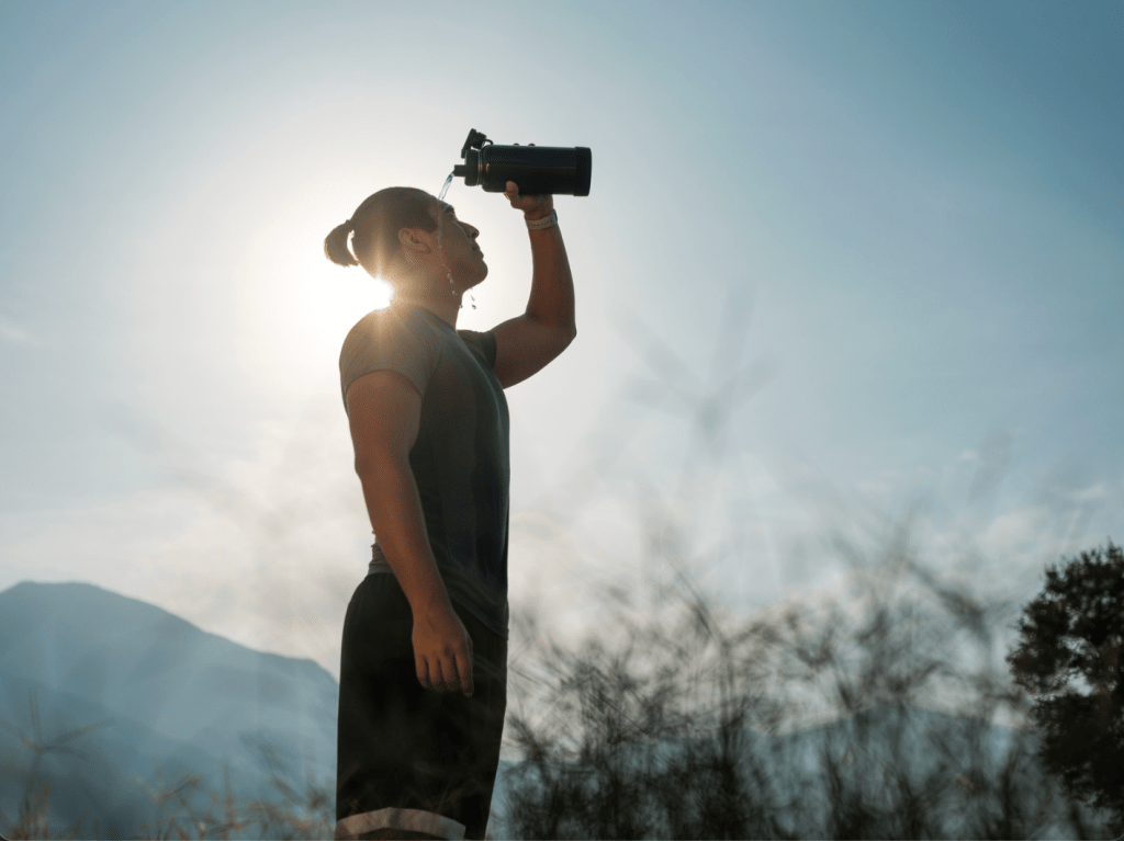 A man in workout clothes standing in a sunny field with mountains inthe background, pours water over his head.