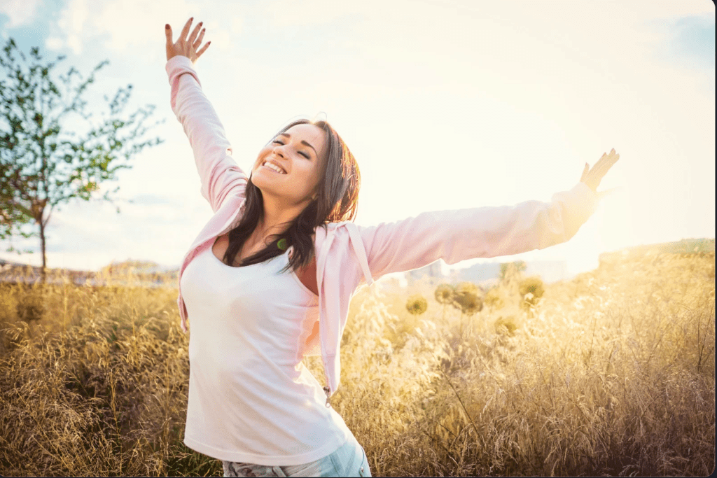 A woman in a sunny field, smiling with her arms extended in the air expressing joy.