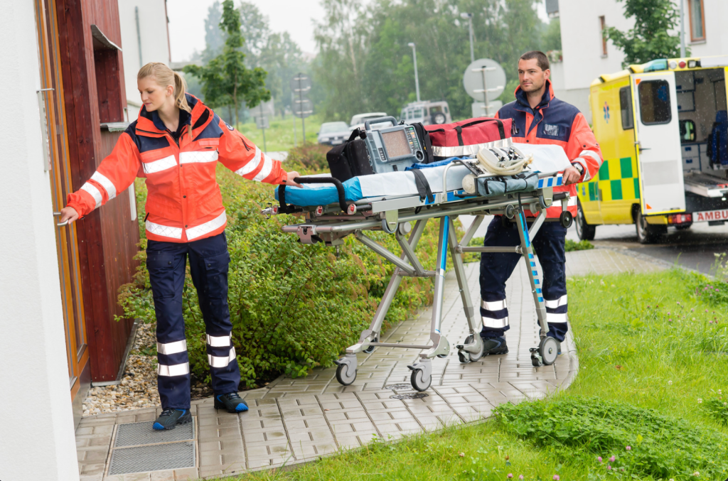 Paramedics wearing orange wheeling a gurney down a path to the front door of a house.