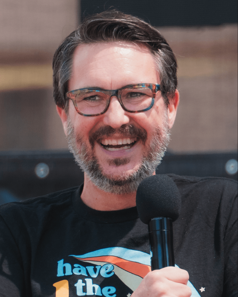 Wil Wheaton smiling and speaking into a microphone, wearing glasses with a salt and pepper beard.