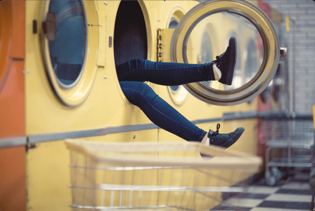 A pair of legs in dark jeans and black high-top sneakers hangs out of an open front-loading washing machine at a laundromat, dangling above an empty laundry basket, with rows of yellow washers and dryers in the background.