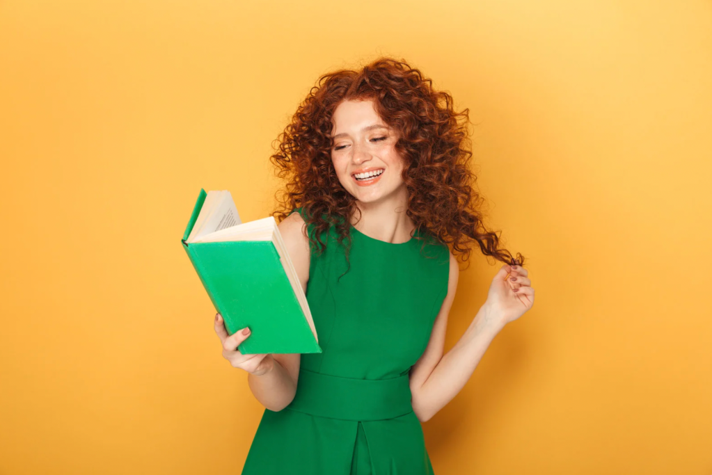 A woman with curly red hair, wearing a sleeveless green dress, smiling while reading a book with a green cover.