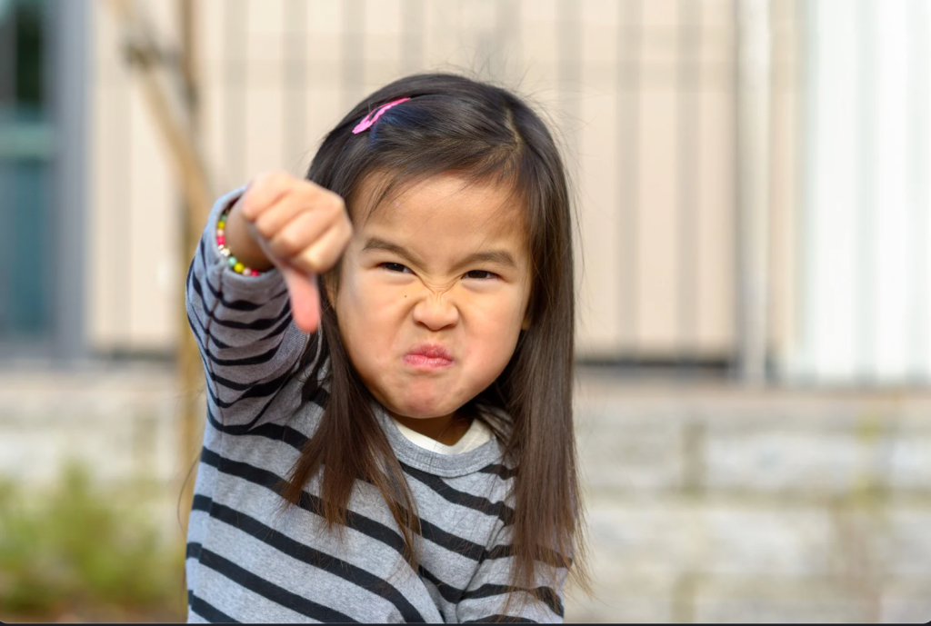 A 5-year-old girl wearing a gray and blue striped sweater giving a thumbs down with an angry expression on her face.