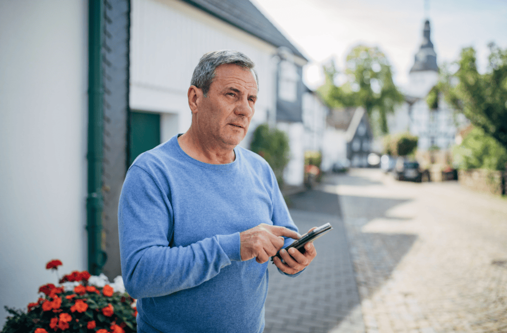 Gray-haired man in a blue sweater stands on a cobblestone street, tapping a smartphone and looking off to the side with a thoughtful expression