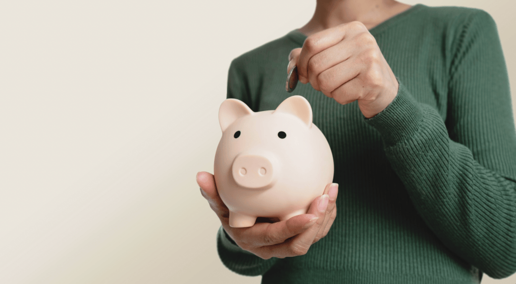 Person in a green sweater holding a pink piggy bank in one hand and dropping a coin into it with the other