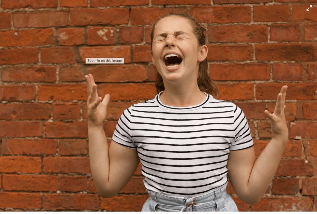 Young woman with eyes closed, mouth open mid-scream or shout, raising both hands against a brick wall background